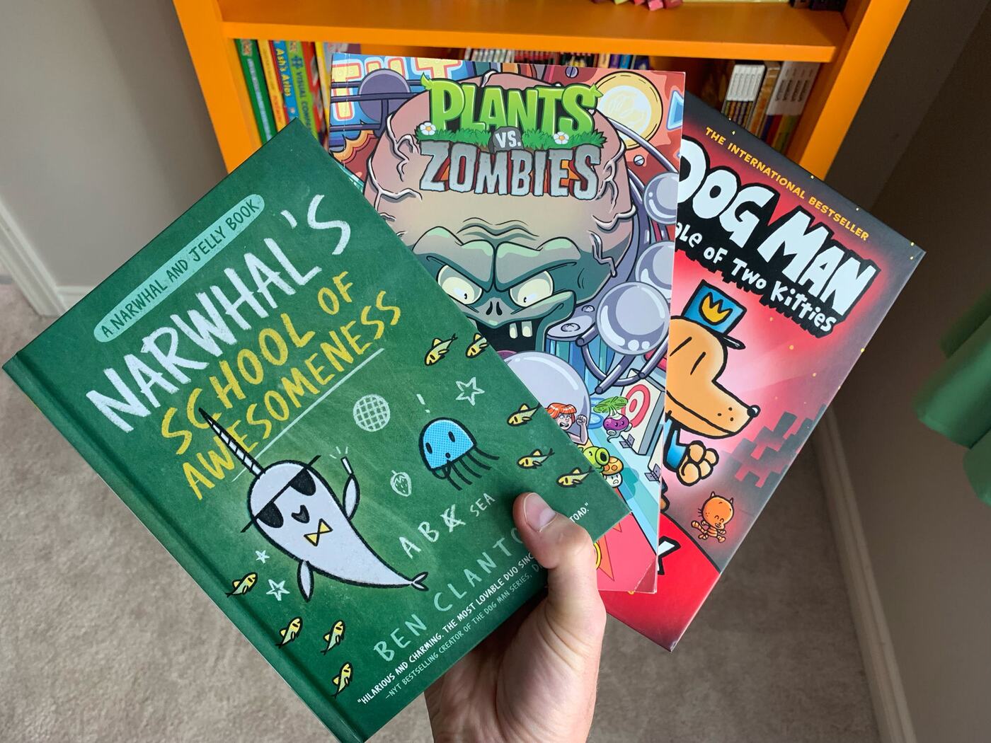 Narwhal, Plants vs. Zombies and Dog Man graphic novels held in front of a bookshelf